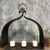 Cast iron triple candle holder with three glass hurricanes and white pillar candles, dark distressed finish, arched top.