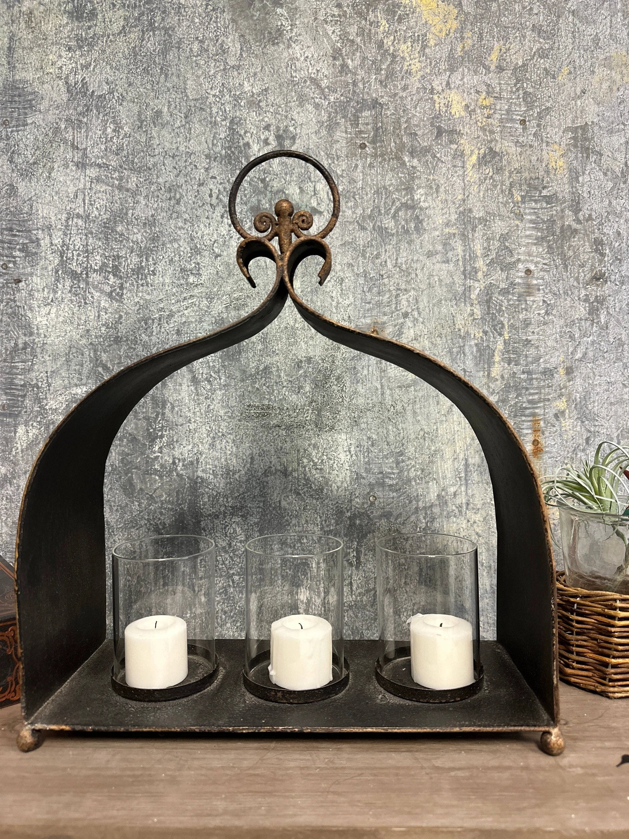 Cast iron triple candle holder with three glass hurricanes and white pillar candles, dark distressed finish, arched top.