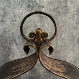 Rusty cast-iron triple candleholder with circular handle and ornate scrollwork against a weathered gray background.