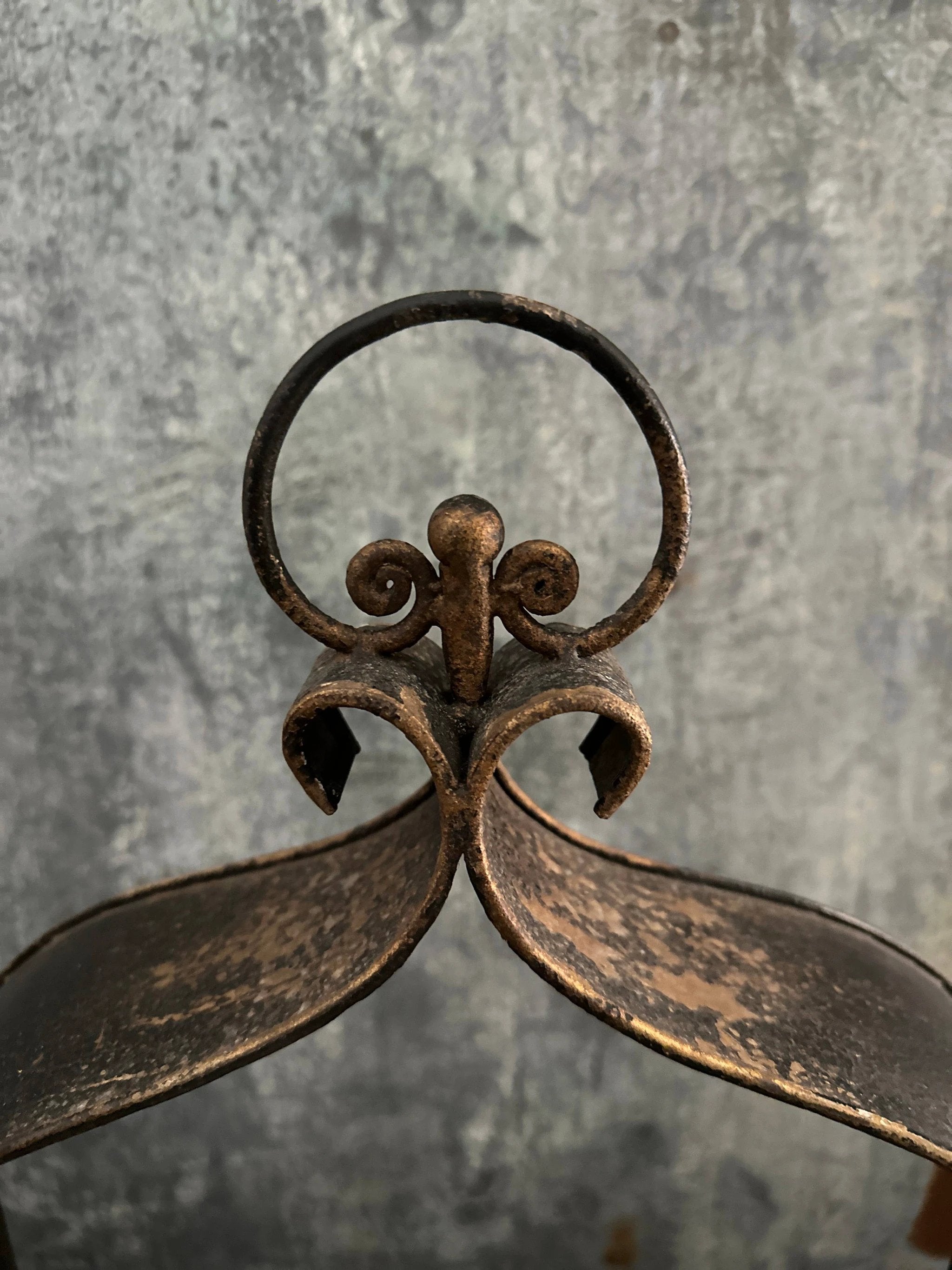 Rusty cast-iron triple candleholder with circular handle and ornate scrollwork against a weathered gray background.