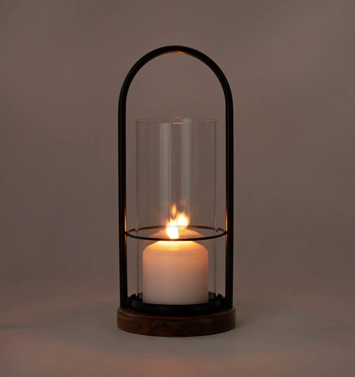 Black metal arch over a clear glass cylinder housing a lit white pillar candle, set on a round wooden base.