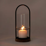 Black metal arch over a clear glass cylinder housing a lit white pillar candle, set on a round wooden base.