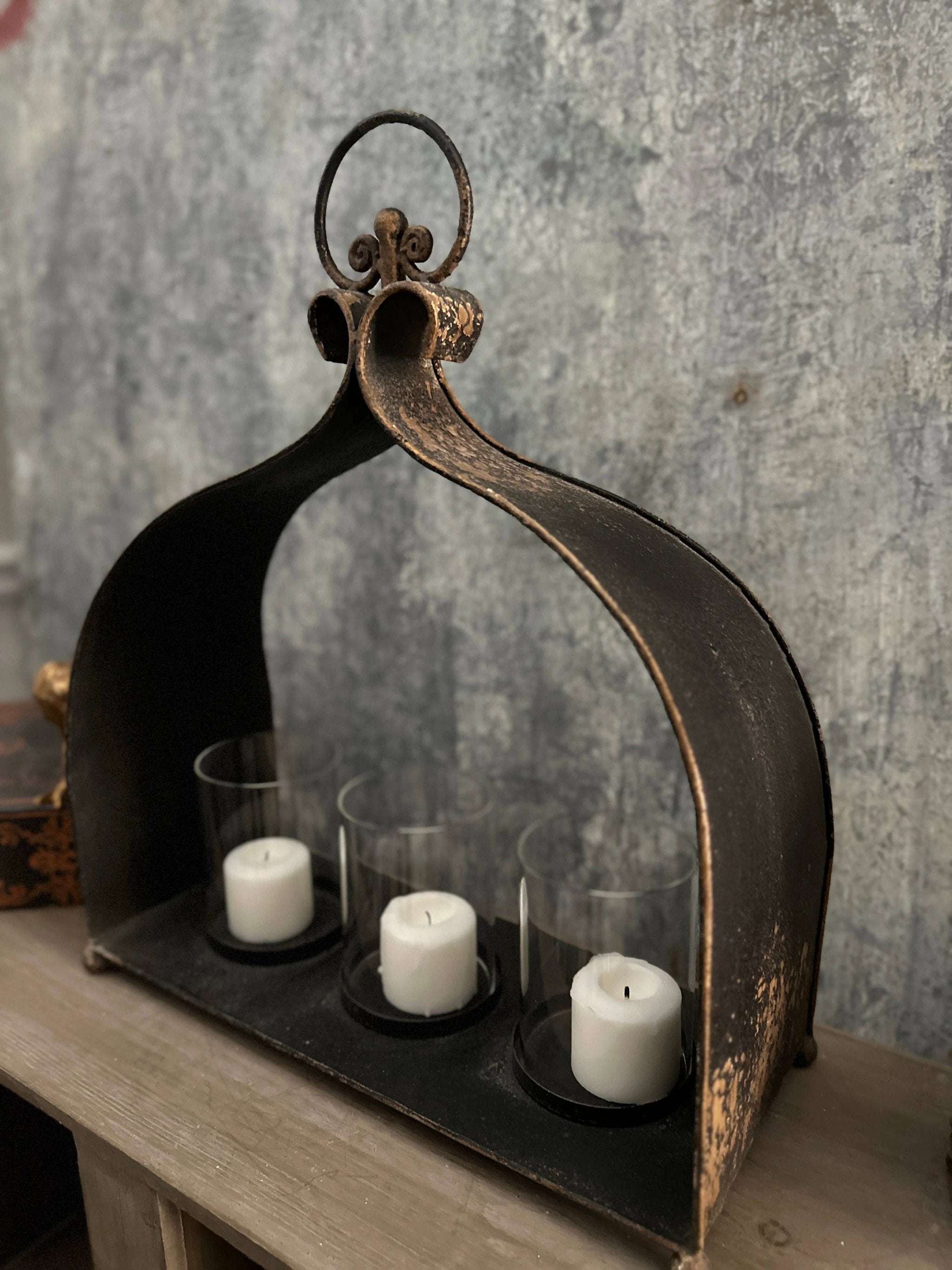 Rustic black cast-iron triple candleholder with three glass cylinders and white pillar candles, arched frame and ring.