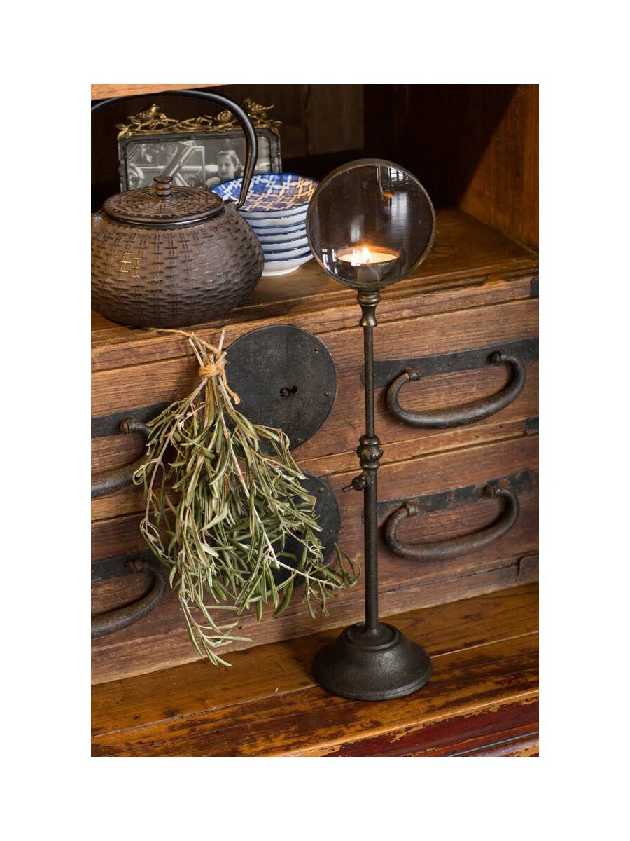 Tall black iron candle holder with a circular magnifying lens at the top; lit candle visible through the lens.
